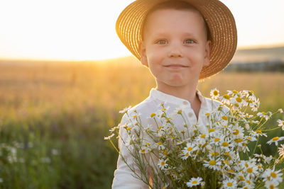 Portrait of smiling boy wearing hat holding flowers on field