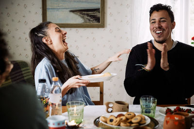 Young woman laughing while looking at male friend clapping hands in house