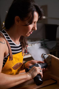 Side view of concentrated adult female carpenter with dark hair and tattoos on arm in casual clothes and apron using drill while screwing metal wheels to wooden board in workshop