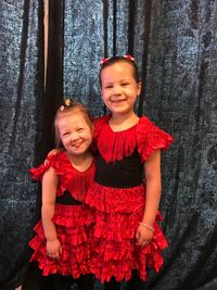 Portrait of smiling sisters in red dress standing against curtain at home