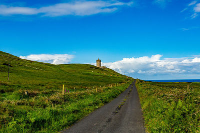 Rural road amidst field against sky, irish countryside with the tower of doonagore castle background