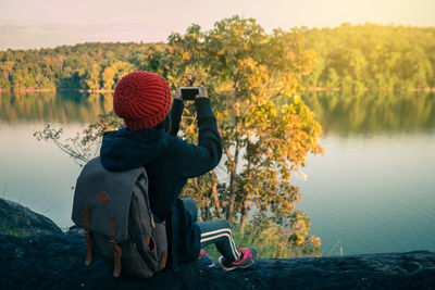 Rear view of girl photographing at lakeshore against trees