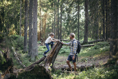 Side view of daughter climbing log by father in forest