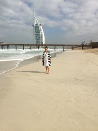 Woman standing at beach with burj al arab hotel in background against cloudy sky