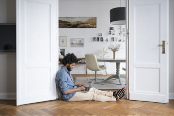Man at home sitting on floor working with laptop in door frame
