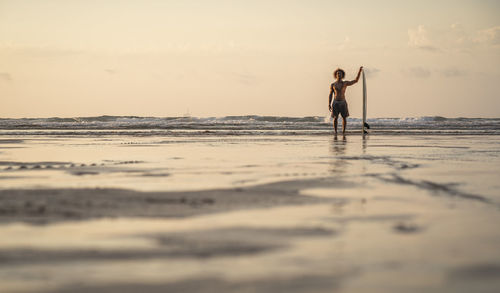 Shirtless man standing with surfboard at seashore against sky during sunset