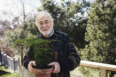 Portrait of senior man holding potted plant while standing against trees