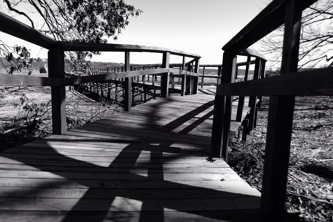 railing, tranquility, shadow, built structure, sunlight, the way forward, tranquil scene, water, wood - material, sky, tree, nature, clear sky, pier, bridge - man made structure, day, scenics, no people, connection, sea