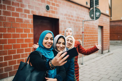 Happy woman taking selfie with female friends on sidewalk against building in city