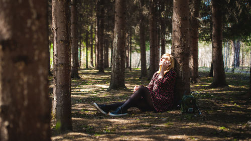 Woman talking on mobile phone while sitting by tree trunk in forest