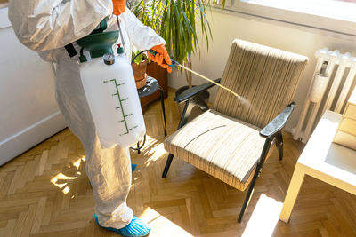 Specialist in protective suit with decontamination sprayer bottle disinfecting household furniture