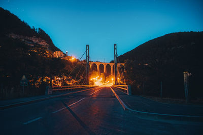 Road by illuminated bridge against sky at night