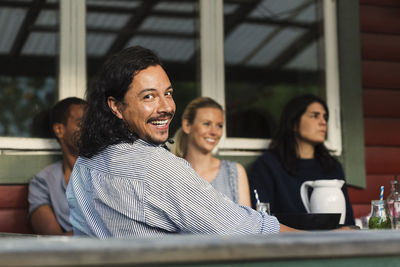 Rear view portrait of happy man sitting with friends at log cabin