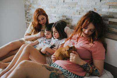 Young same gender parents and kids in pajamas playing with guinea pigs and relaxing on bed in bedroom