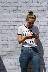 Smiling teenage girl using mobile phone while standing against brick wall