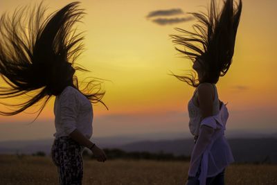 Side view of women tossing hair while standing on land against sky during sunset