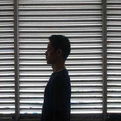 Side view of young man standing by window blinds