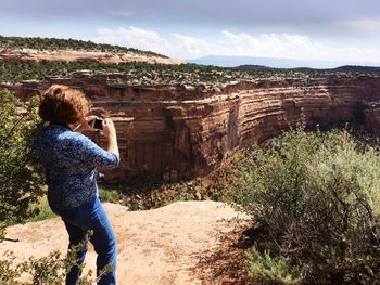Side view of woman photographing rock formation against sky