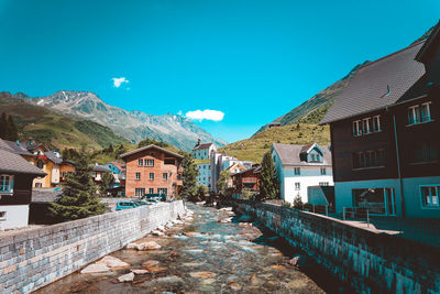 Canal flowing amidst buildings by mountains against blue sky