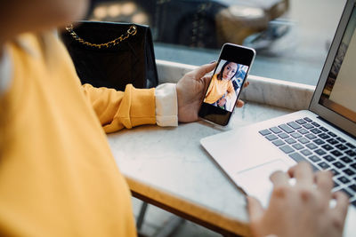 Midsection of female freelancer holding smart phone using laptop at cafe