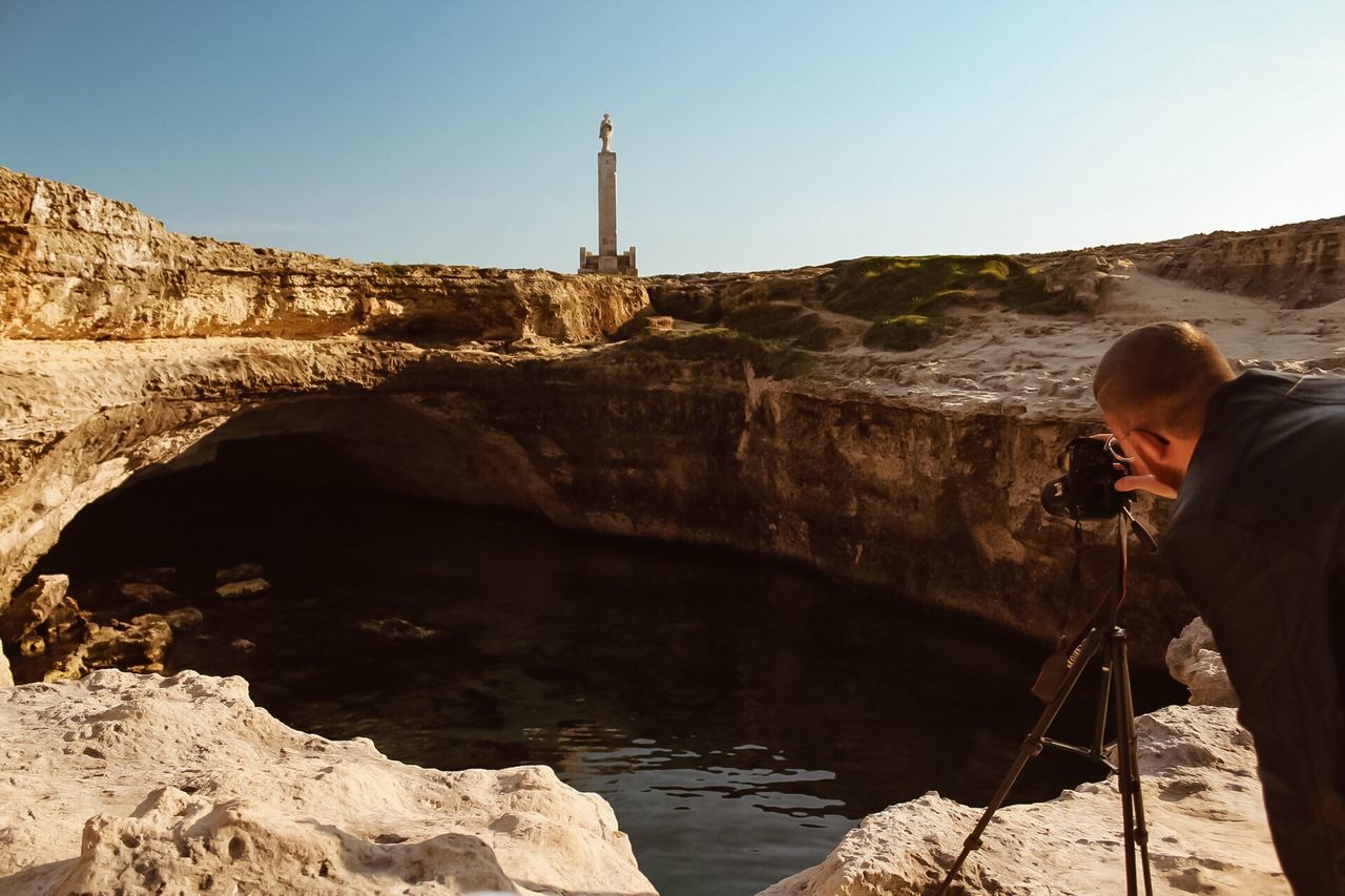 MAN PHOTOGRAPHING ON ROCK BY WATER