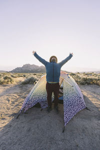 Full length of tourist stretching arms while standing by tent at desert against clear sky during sunrise