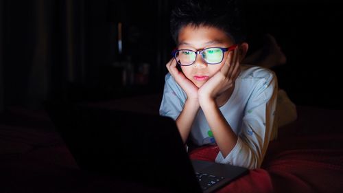 Boy using laptop while lying on bed in darkroom at home