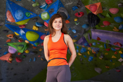 Charming strong woman in sportswear holding hands behind back while standing against climbing wall and looking at camera