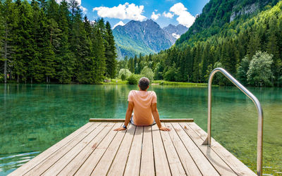 Rear view of young man sitting on deck by lake in mountains.