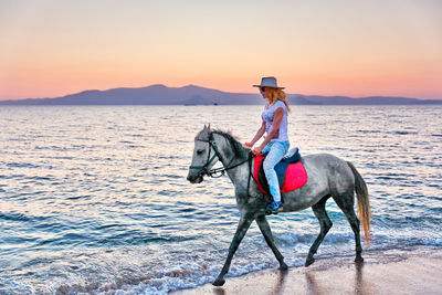 Boy riding at shore against sky during sunset