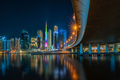 Illuminated modern buildings by river against sky at night