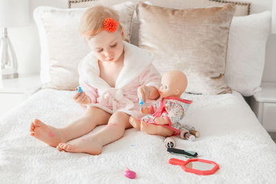 Cute girl applying make-up to doll while sitting on bed at home