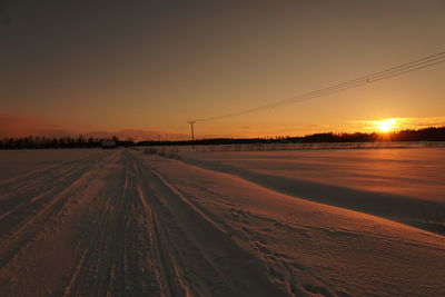 Tire tracks on snow covered land against sky during sunset