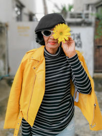 Senior woman with jacket wearing sunglasses holding yellow flower in city