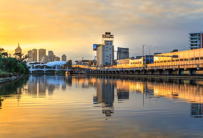 Bridge over river by buildings against sky during sunset