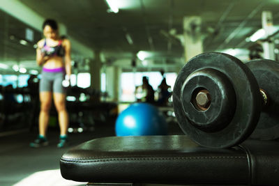 Close-up of dumbbell on seat with woman exercising at gym