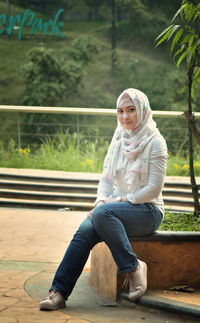 Portrait of smiling young woman sitting on retaining wall at park