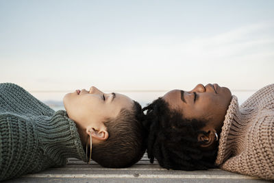 Female friends with eyes closed lying on bench against sky