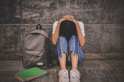 Depressed female student sitting with books and backpack against wall