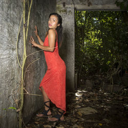 Side view of woman wearing red dress while standing by wall in forest