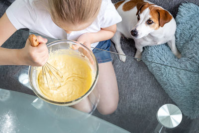 A girl stirs the dough with a whisk in a glass bowl, and a hungry dog sits nearby. cook at home.