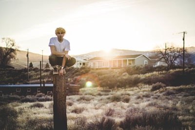 Portrait of man crouching on wooden post against sky during sunset