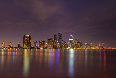 Illuminated buildings by river against sky at night