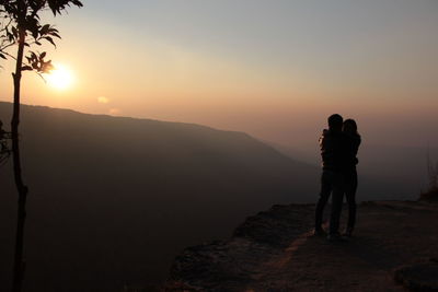 Couple standing on mountain against sky during sunset