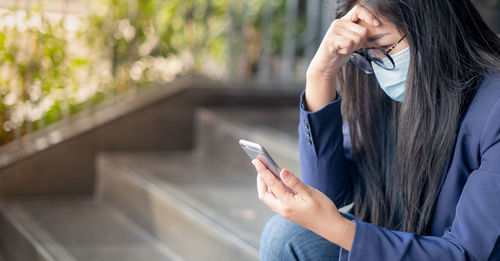Depressed businesswoman wearing mask using mobile phone outdoors
