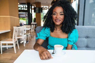 Content african american lady with makeup sitting at table with cup of coffee and looking at camera in cafe