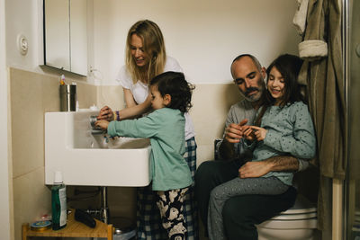 Woman helping son with washing hands while man and girl talking in bathroom