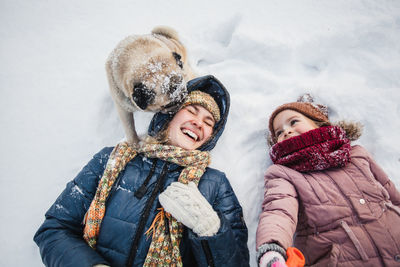 High angle view of dog licking cheerful mother lying with daughter on snow