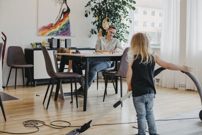 Mother sitting at table while girl hoovering