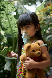 Girl breathing with nebulizer while standing with teddy bear by plants